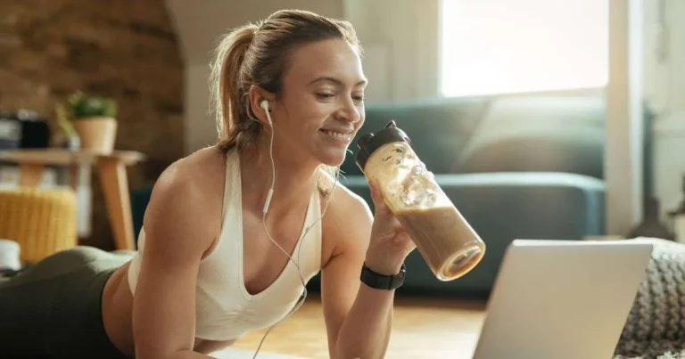 Mulher com roupas de treino tomando shake proteico em casa enquanto assiste a conteúdo no notebook.
