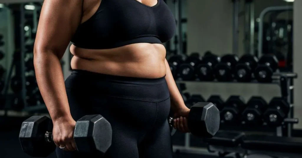 Mulher plus size segurando halteres em academia, com foco no corpo e no esforço durante o treino de musculação