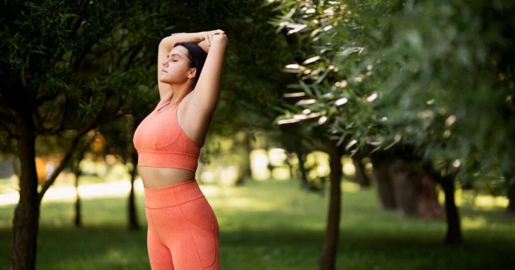 Mulher com roupas de treino alongando os braços acima da cabeça em parque arborizado, com olhos fechados e postura relaxada