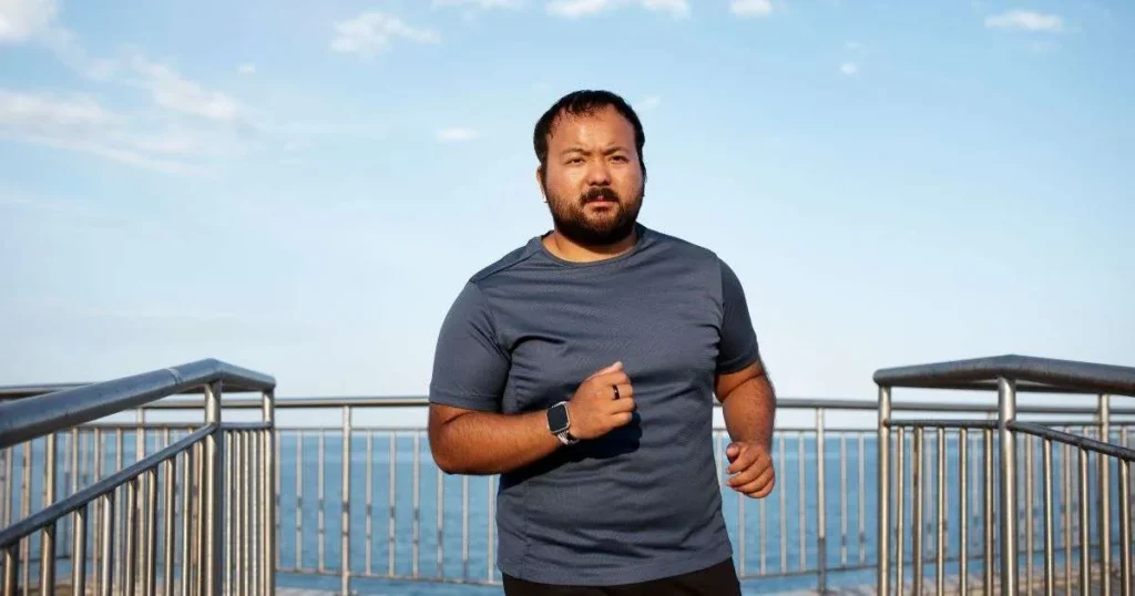 Homem com sobrepeso praticando corrida ao ar livre em um calçadão próximo ao mar, representando o esforço físico no combate à obesidade e na regulação dos hormônios masculinos.