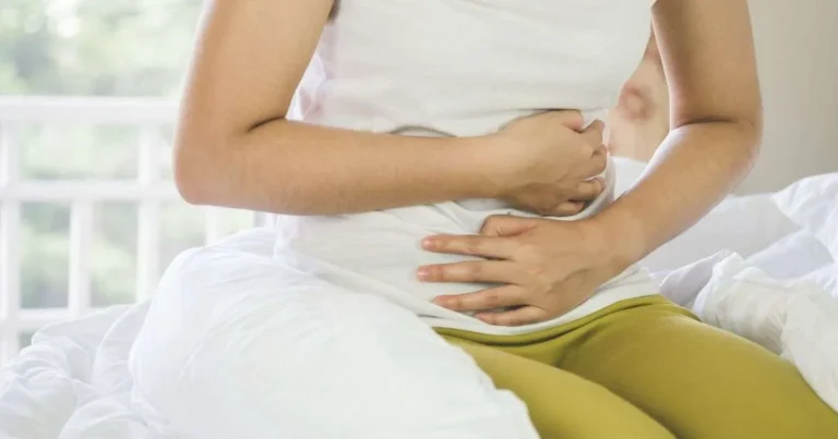 Constipação intestinal. Pessoa sentada na cama segurando o abdômen com as duas mãos, demonstrando desconforto abdominal