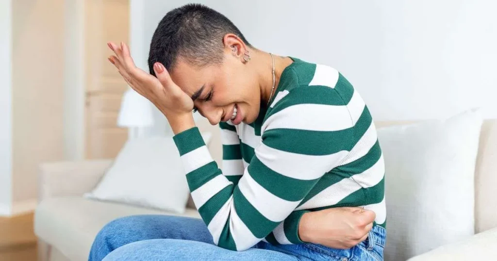 Pessoa jovem sentada no sofá curvada para frente com expressão de dor, segurando o abdômen e a cabeça simultaneamente
