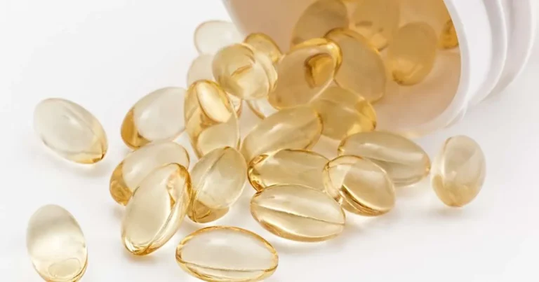 Cápsulas gelatinosas douradas de vitamina d espalhadas ao lado de um frasco branco sobre fundo claro