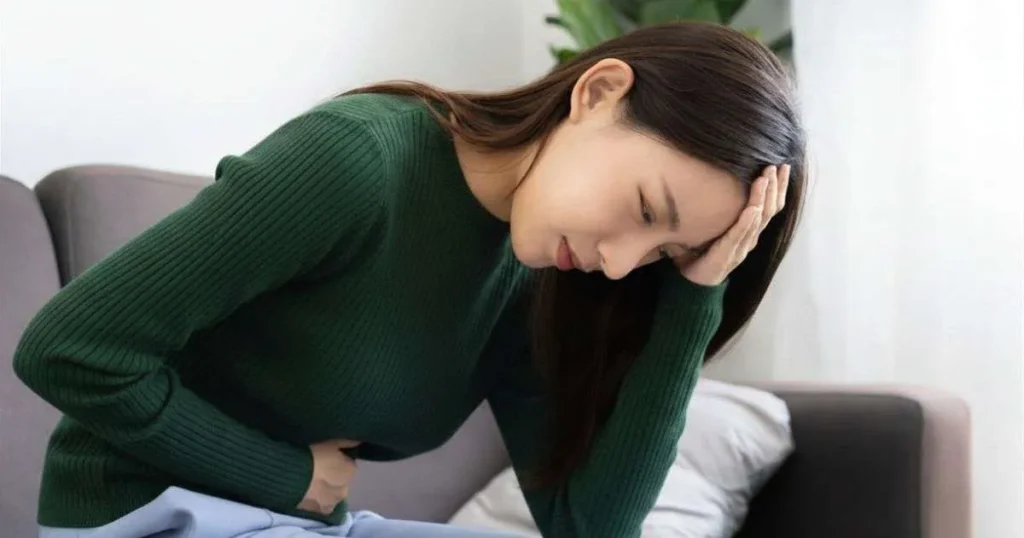 Mulher jovem curvada no sofá com expressão de dor, segurando o abdômen com uma mão e apoiando a cabeça com a outra
