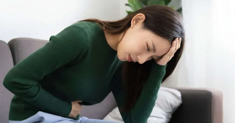 Mulher jovem curvada no sofá com expressão de dor, segurando o abdômen com uma mão e apoiando a cabeça com a outra
