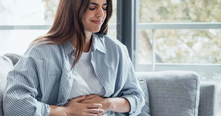 Mulher sentada no sofá segurando o abdômen com expressão serena, sugerindo desconforto leve ou atenção à região intestinal