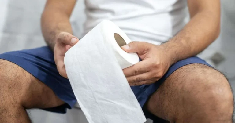 Homem sentado segurando rolo de papel higiênico, sugerindo dificuldade ou demora no banheiro por constipação