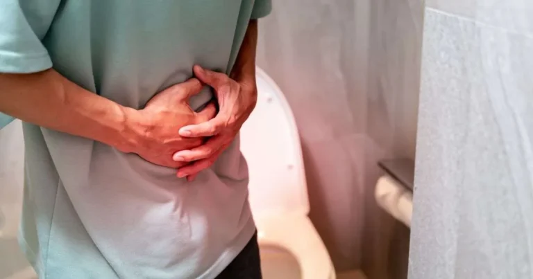 Pessoa curvada segurando o abdômen com as mãos próxima ao vaso sanitário, demonstrando cólica ou urgência intestinal