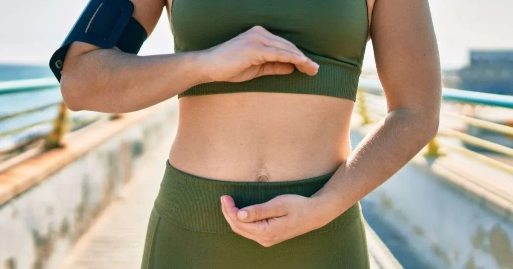 Mulher com roupa de treino enquadrando o abdômen com as mãos ao ar livre, com foco na região intestinal