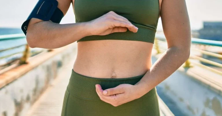 Mulher com roupa de treino enquadrando o abdômen com as mãos ao ar livre, com foco na região intestinal