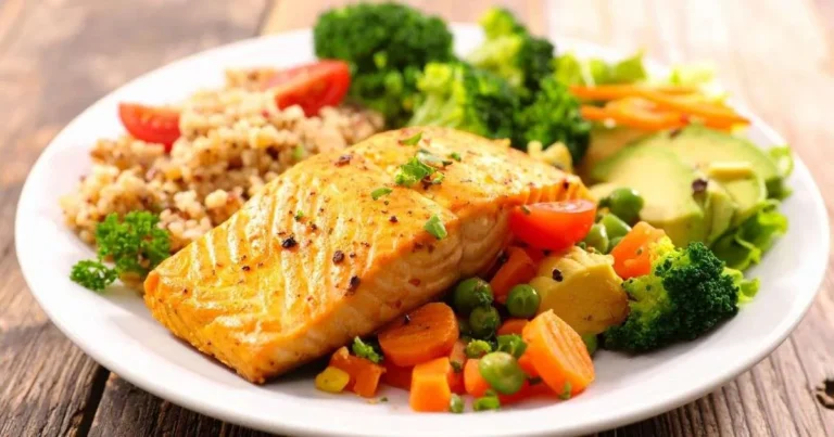 Prato com filé de salmão grelhado, brócolis, cenoura, ervilha, abacate e quinoa sobre mesa de madeira