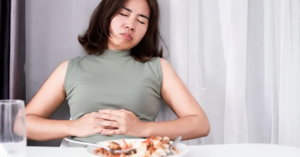 Mulher à mesa com prato de comida à frente segurando o abdômen com expressão de desconforto após comer