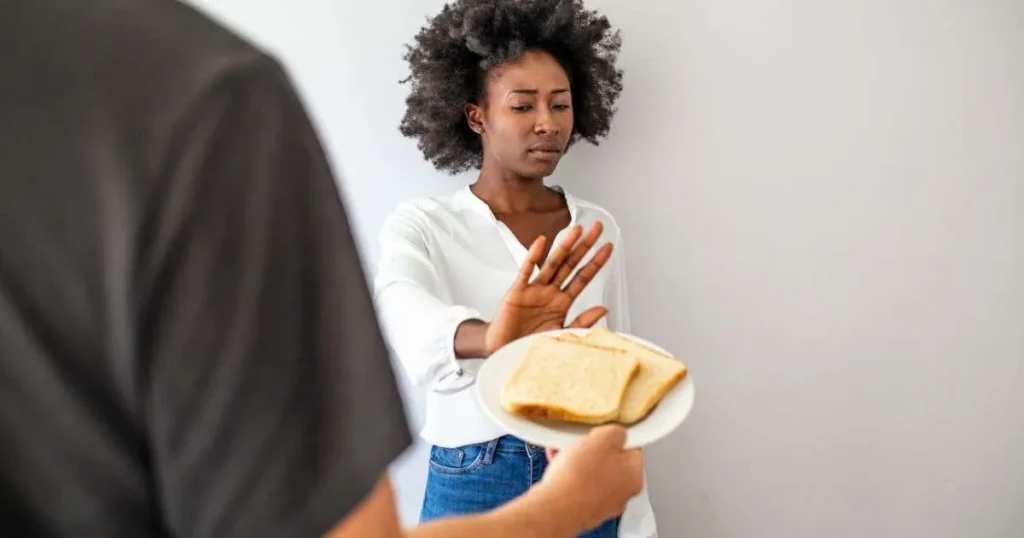 Mulher recusando pão oferecido em prato com gesto de negação e expressão de desconforto