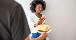 Mulher recusando pão oferecido em prato com gesto de negação e expressão de desconforto