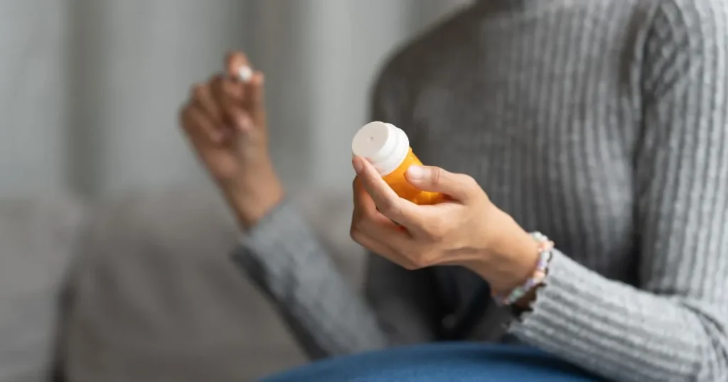 Pessoa segurando frasco laranja de suplemento e cápsula na outra mão, prestes a ingerir o medicamento