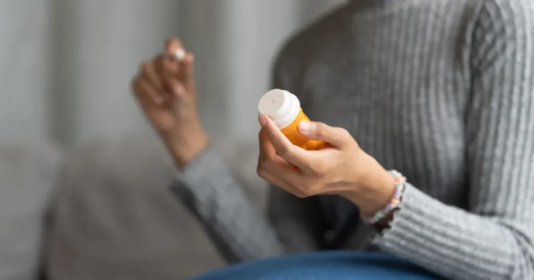 Pessoa segurando frasco laranja de suplemento e cápsula na outra mão, prestes a ingerir o medicamento